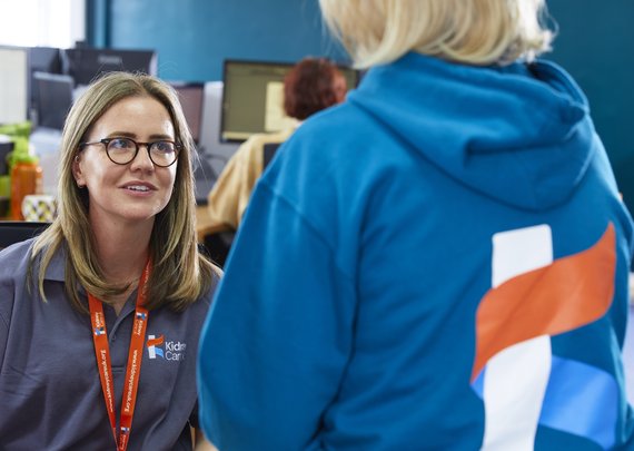 Kidney Care UK staff member, wearing a branded polo shirt and lanyard, chatting at her desk with another member of staff.