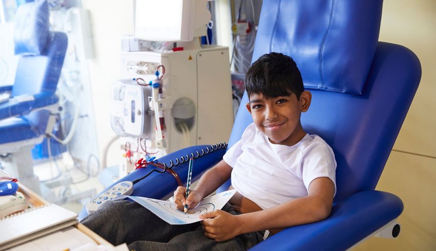 Boy in a hospital bed drawing on a pad of paper. Next to him is a haemodialysis machine. He has a line for vascular access.