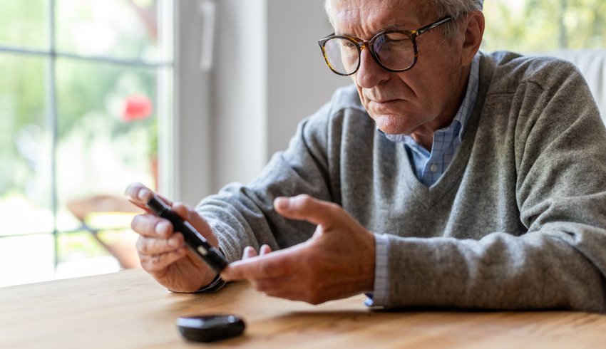 Old man sitting at a table doing a blood sugar pinprick test for diabetes.