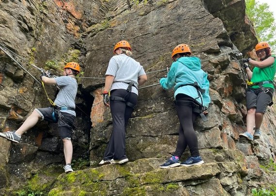 Four young adults, wearing helmets and safety equipment, climing across a rocky outcrop.