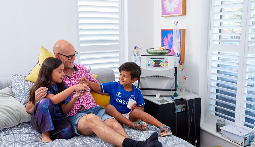 Nick lies on his bed with his two young children, who are eating yoghurt, with his home haemodialysis set up nearby