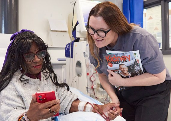 Kidney Care UK Patient Support & Advocacy officer, chatting with a patient who is receiving haemodialysis in a hospital unit. They are both looking at something on the patient's mobile phone.