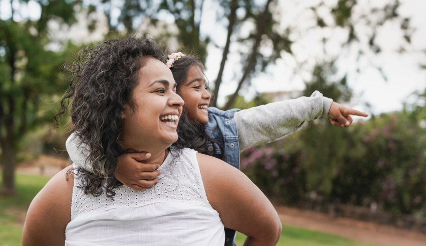 A woman giving a child a piggyback ride in a park, the child is pointing at something